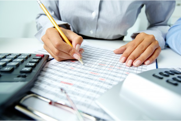 mulher de camisa cinza sentada a uma mesa com uma calculadora faz anotações em uma planilha de papel