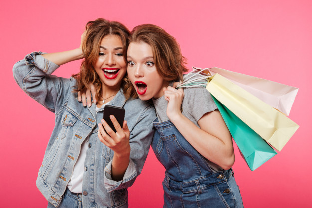 duas amigas com compras se impressionam ao olhar para um celular de costas para um fundo rosa