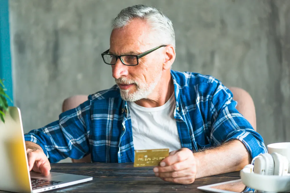 homem de idade mexendo no notebook enquanto segura cartão de crédito consignado