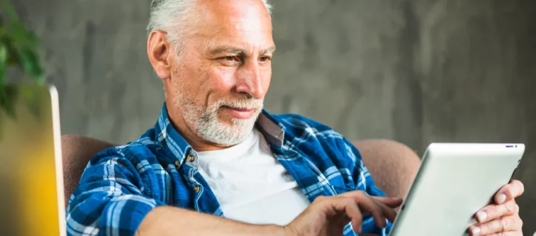 homem de idade mexendo no tablet enquanto sentado ao computador - ate quantos anos aposentado pode fazer emprestimo