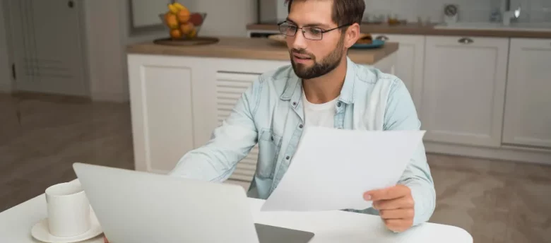homem de óculos sentado na sala de casa com papel na mão em frente ao notebook qual melhor banco para empréstimo consignado inss