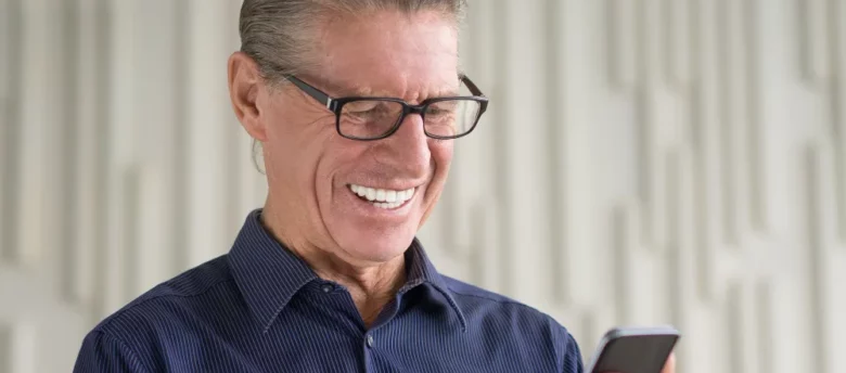 homem de terceira idade usando óculos e sorrindo ao usar o celular - financiamento para aposentado é mais barato