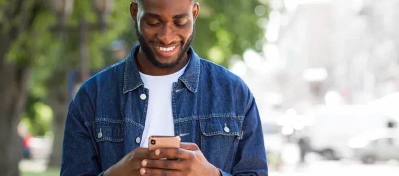 homem negro em pé ao ar livre sorrindo ao olhar o celular na mão - quem faz empréstimo com garantia de celular