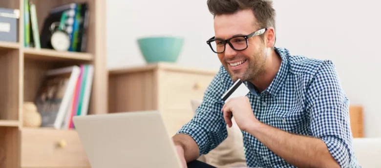 homem sorridente mexendo no notebook e segurando cartão - cartão de crédito para negativado com limite aprovado na hora