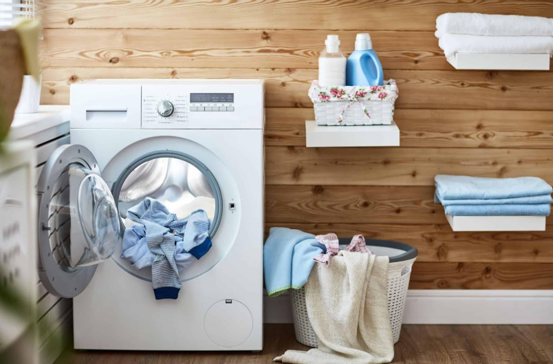 maquina de lavar cheia de roupas dentro de uma lavanderia com parede de madeira e estantes para panos e produtos de limpeza