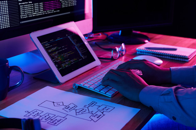 imagem ampliada de desenvolvedor de software trabalhando em uma mesa com duas telas junto de um teclado e uma folha com anotações