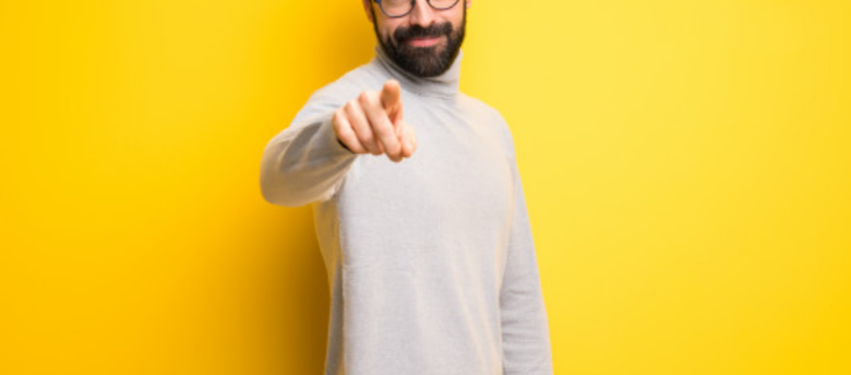homem de camiseta cinza de costas para uma parede amarela aponta para a frente
