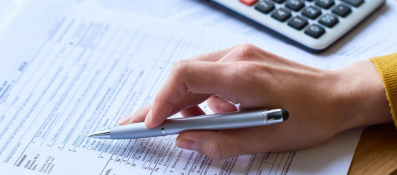 mulher segurando caneta e checando anotações e valores em folha de papel fazendo uso de uma calculadora