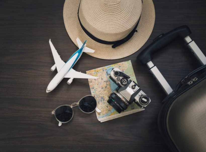 imagem de cima de uma mesa com uma miniatura de aviãol, um chapéu, uma câmera fotográfica, um óculos, um mapa e uma mala de viagens