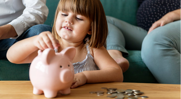 criança pequena sentada ao chão colocando uma moeda dentro de um cofre em formato de porco na cor rosa