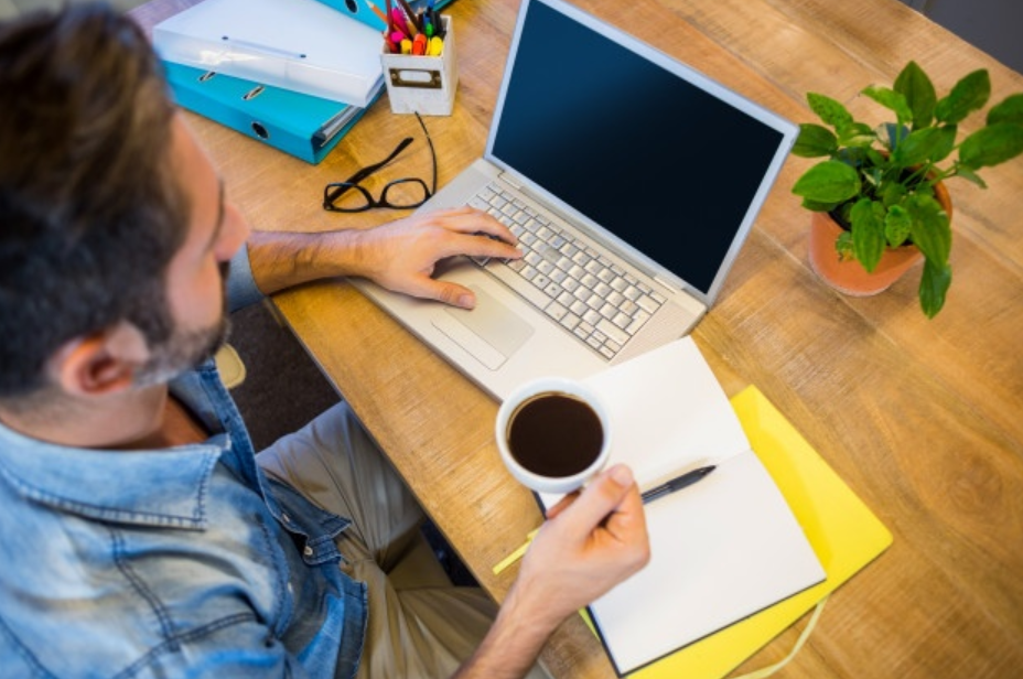 jovem de camisa jeans azul usando seu laptop e bebendo uma xícara de café