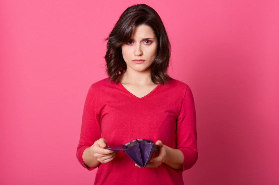 imagem de uma mulher com semblante triste abrindo sua carteira vazia em frente a uma parede rosa