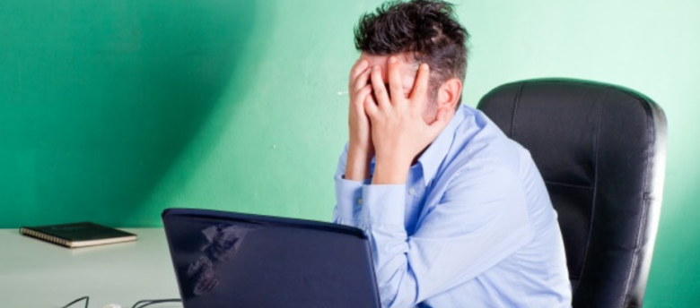 homem de camisa azul sentado em frente a laptop com semblante preocupado e cabeça apoiada nas mãos