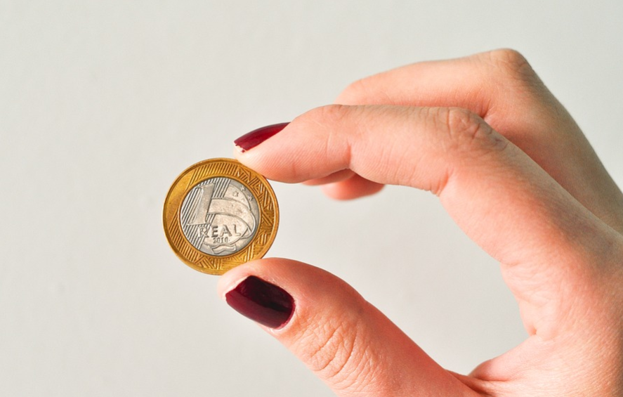 mão com as unhas pintadas segurando uma moeda de 1 real que simboliza poucas taxações dos bancos