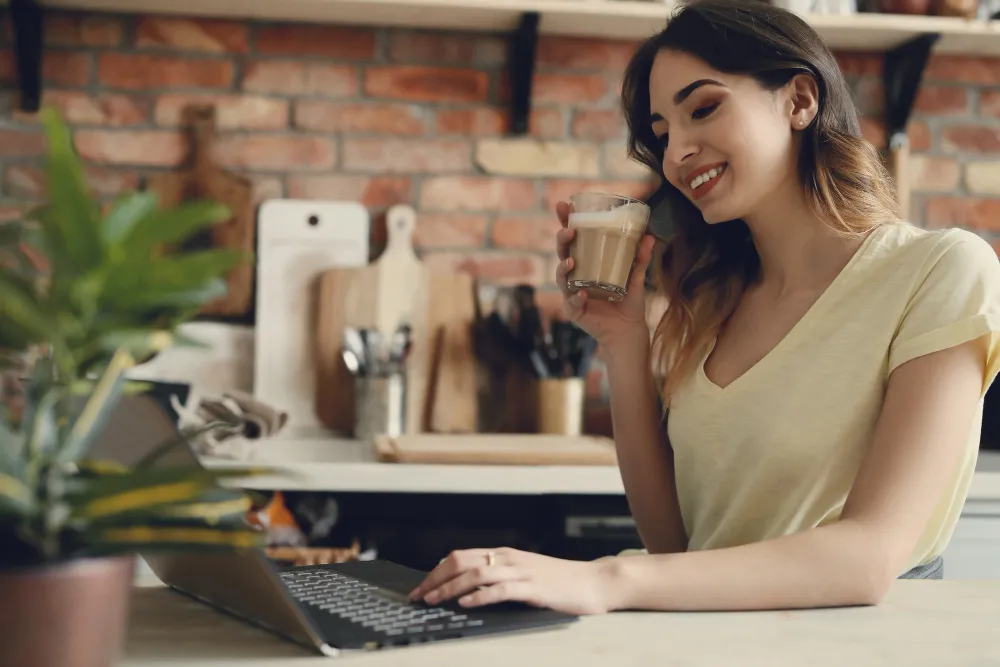 mulher branca sentada à mesa em frente ao computador com copo de café na mão - empréstimo pessoal online seguro e rápido