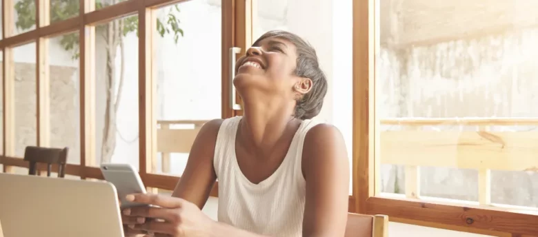 mulher de cabelos curtos sorrindo com o celular nas mãos em frente ao computador - melhor banco para antecipar fgts