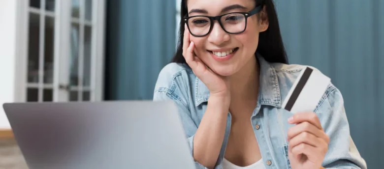 mulher feliz com cartão na mão, sentada em frente ao computador - cartão de crédito para negativado e com score baixo