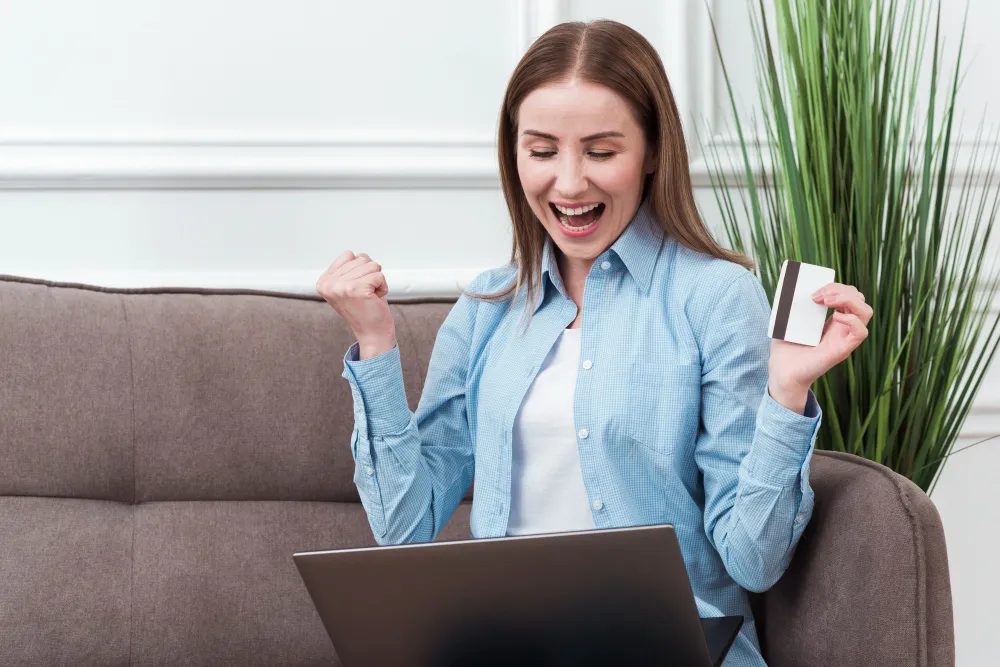 mulher feliz sentada no sofá segurando cartão em frente ao computador - cartão de crédito para negativado e com score baixo