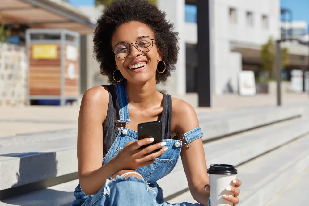 mulher negra ao ar livre sorrindo com café e celular na mão - empréstimo de 300 reais na hora via pix