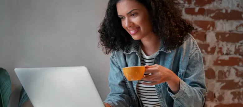 mulher negra sentada em frente ao computador com xícara de café na mão - empréstimo pessoal online seguro e rápido