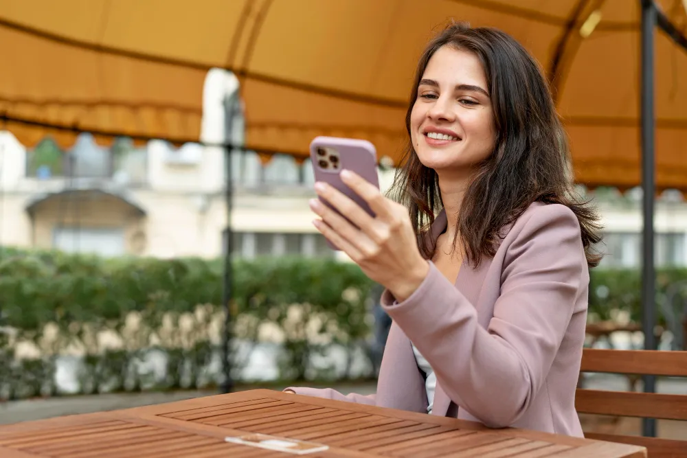 mulher sentada em mesa de madeira na rua sorrindo com celular na mão - quem faz empréstimo com garantia de celular