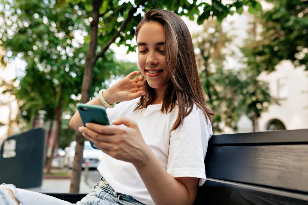 mulher sorridente sentada em banco de praça mexendo no celular - calendário bolsa família 2024