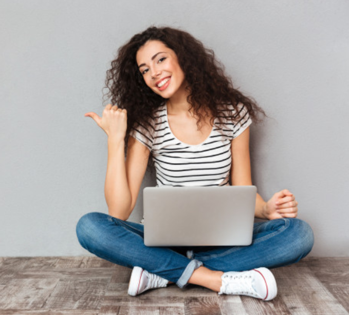 mulher sentada ao chão vestindo camiseta listrada e calça jeans e usando um laptop