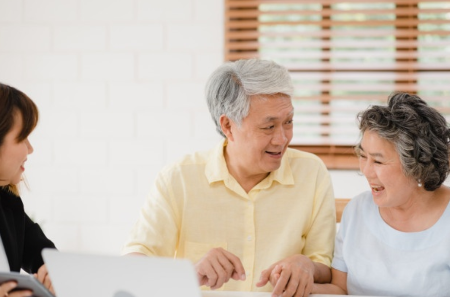 casal de idosos sendo ele de camisa amarela e ela de blusa branca conversando com bancária de terno para entender as novas regras do empréstimo consigando