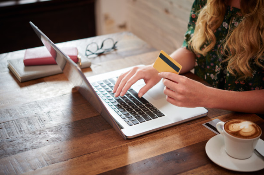 imagem ampliada de mulher sentada a uma mesa usando um laptop e segurando um cartão de crédito enquanto toma um café