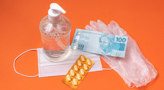 em uma mesa laranja estáum frasco com álcool em gel, uma máscara cirúrgica, luvas, comprimidos e notas de cem reais