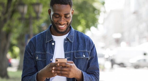 homem de jaqueta jeans parada em calçada sorrindo enquanto usa o celular