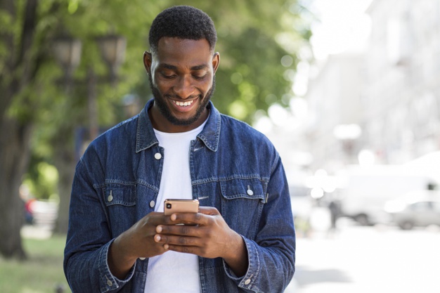 homem de jaqueta jeans parada em calçada sorrindo enquanto usa o celular