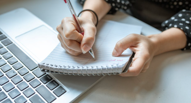 imagem ampliada da mão de mulher escrevendo em caderninho em frente a computador