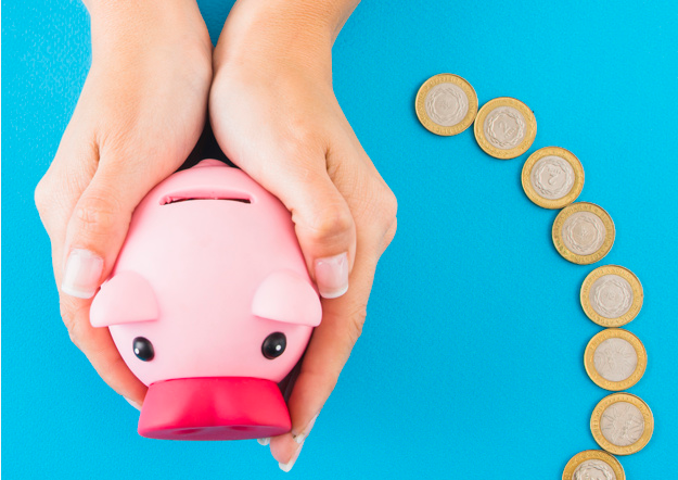 pessoa segurando um cofre em formato de porco rosa em uma mesa azul rodeada de moedas de um real