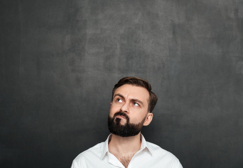 homem de camisa branca usando barba de pé à frente de fundo cor grafite pensando sobre como solicitar o interveniente auitante