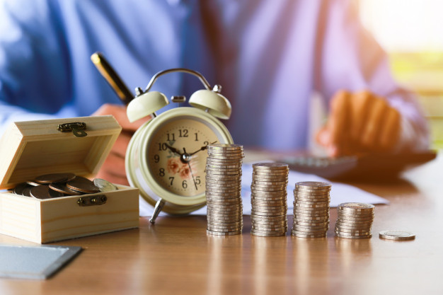 moedas e relógio despertador sobre mesa em que homem escreve em uma folha e faz contas na calculadora