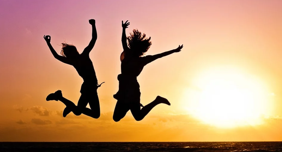 imagem do por do sol com a silhueta de duas mulheres que estão pulando e bem alegres. O pulo delas congelou no ar