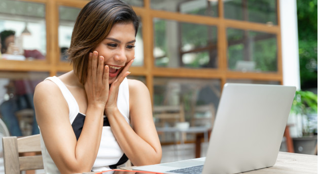 mulher sorrindo e levando as mãos ao rosto sentada de frente para um laptop