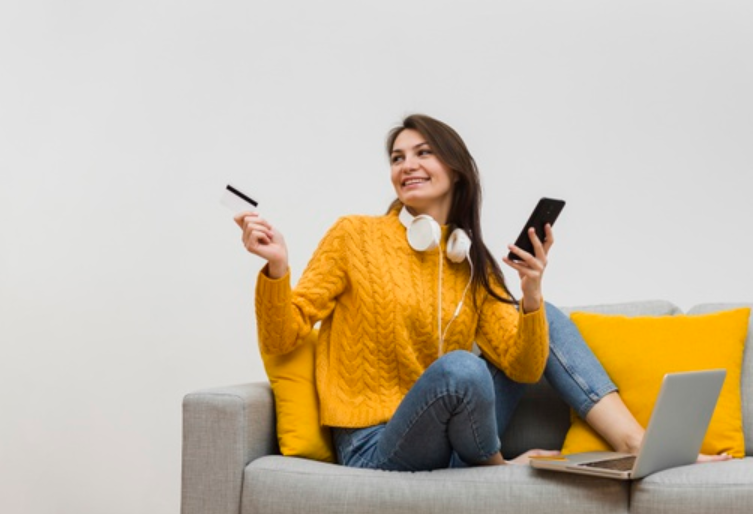 mulher de blusa amarela e calça azul escutando música e sentada em um sofá segurando um cartão de crédito