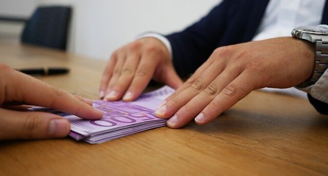 Empresário empurrando dinheiro com as duas mãos para um moço sentado à frente da mesa