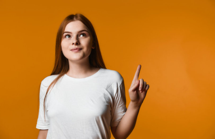 garota ruiva de camiseta branca com semblante pensativo em frente a uma parede laranja