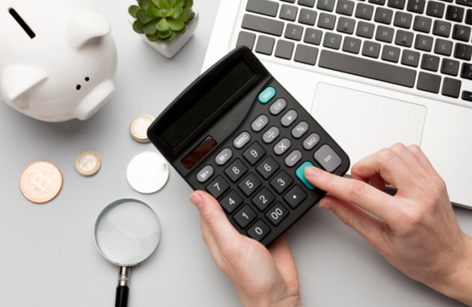 imagem ampliada de uma pessoa usando a calculadora em cima de uma mesa com um laptop, um cofre, uma lupa e moedas