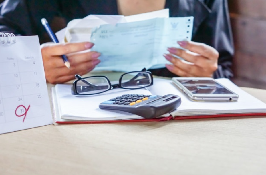 imagem aproximada de pessoa sentada a uma mesa com contas, óculos, caderno, calculadora e um celular