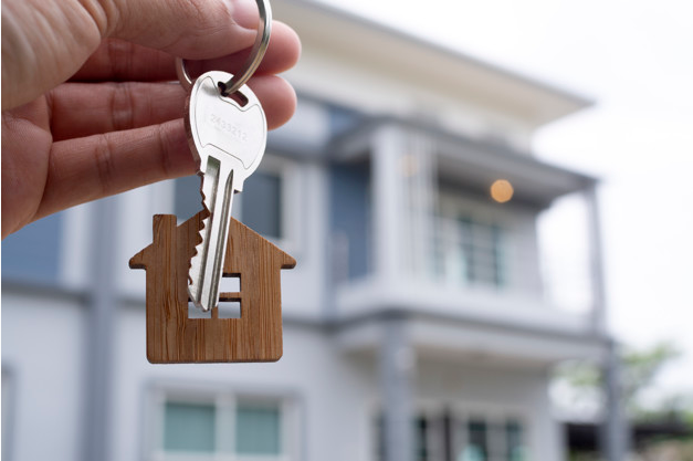 imagem ampliada em primeira plano a mão de uma pessoa segurando um chaveiro com uma chave e uma casa de madeira em miniatura e ao fundo imóvel de dois andares na cor branca