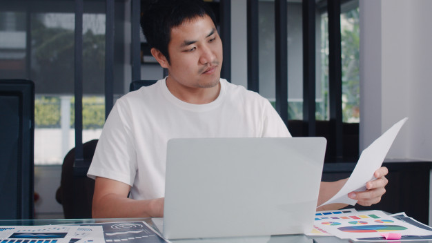 homem de camiseta branca sentado em frente a um laptop olhando para uma folha de papel