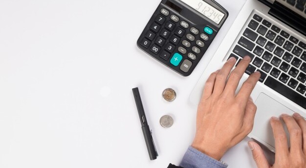 mesa branca com mãos usando um laptop, uma calculadora, moedas e uma caneta