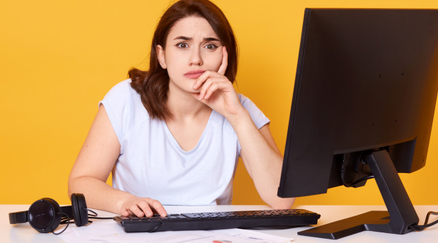 mulher camiseta branca com semblante preocupado sentada a uma mesa com um monitor e um teclado de costas para um fundo amarelo