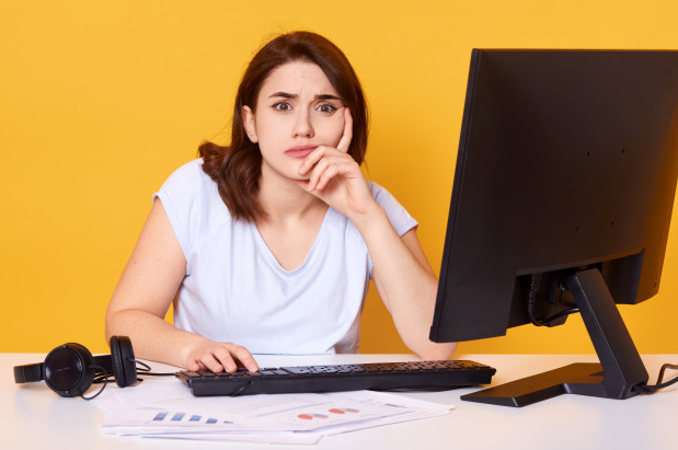 mulher camiseta branca com semblante preocupado sentada a uma mesa com um monitor e um teclado de costas para um fundo amarelo