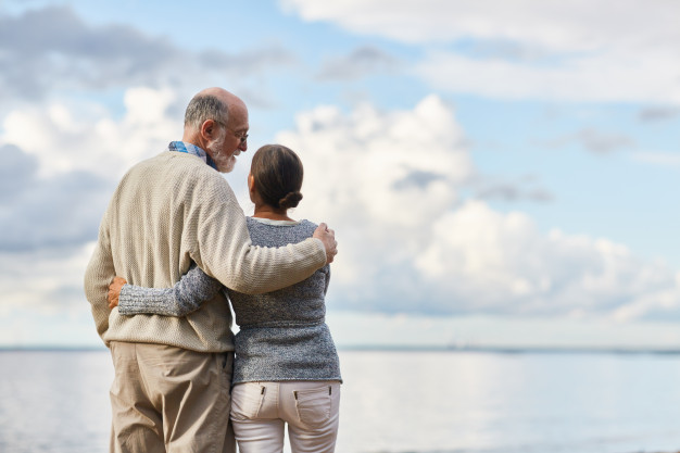 imagem de casal de idosos de costas se abraçando próximos ao mar