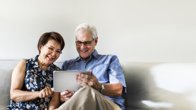 casal idoso sentado em sofá sorrindo e usando um tablet